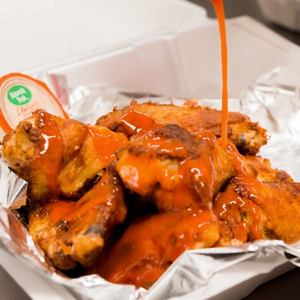Buffalo sauce being poured over a container of crispy chicken wings, with a side of ranch.