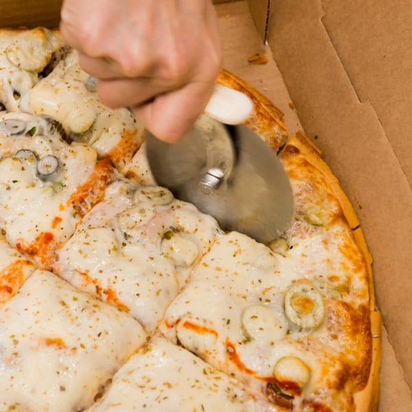A hand uses a pizza cutter to slice a Bob's Tu Your Door Pizza with melted cheese and toppings.